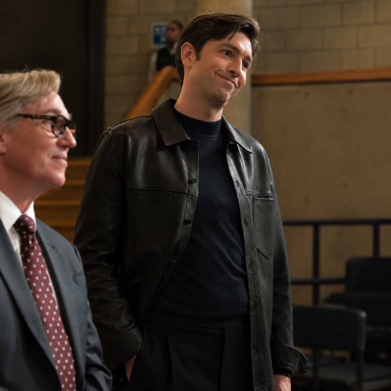 Jeremy-perrick-elsbeth-season-4-leather-trucker-jacket Man wearing a black leather trucker jacket over a black shirt in a cinematic indoor setting.