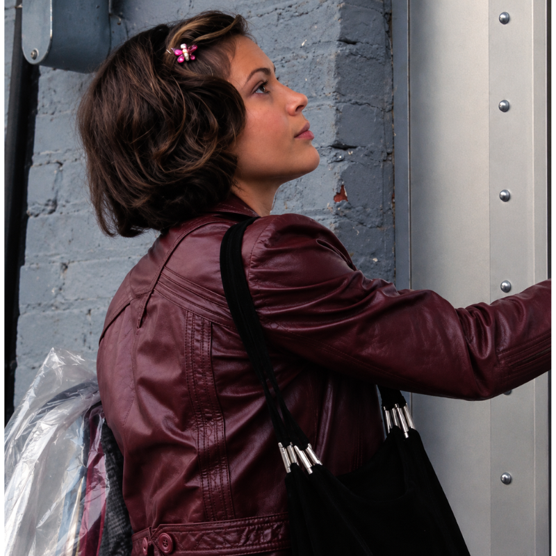 Phoebe-halliwell-charmed-1998-season-1-burgundy-jacket Woman wearing a burgundy leather-look blazer jacket with tailored seams and a waist strap beside a grey industrial doorway.
