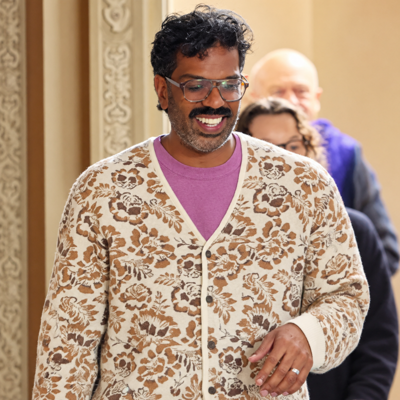 Romesh-ranganathan-cream-floral-jacquard-cardigan Model wearing a cream floral jacquard cardigan over a purple shirt, showing the V-neck, dark brown buttons, and long sleeves.