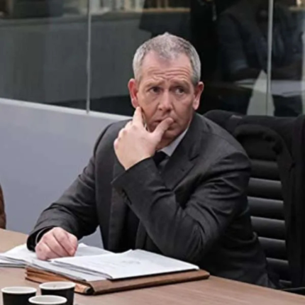 Man wearing a black business suit and tie while sitting at a conference table with documents and a coffee cup.