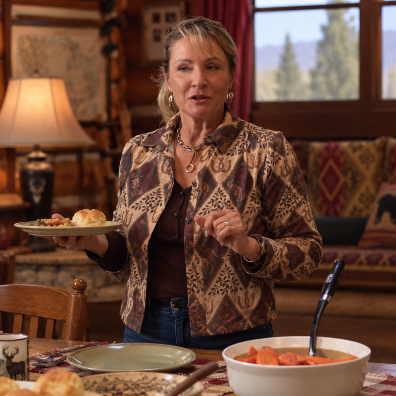 lisa-stillman-heartland-season-20-jacket Woman wearing a tan and brown Western printed jacket with rust-red geometric floral motifs over a dark top and blue jeans in a rustic indoor setting.